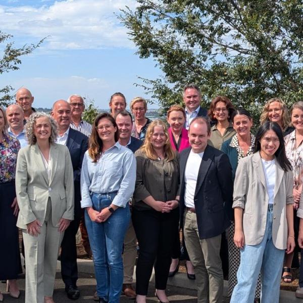 A group of colleagues stand under blue skies and trees, smiling at the camera