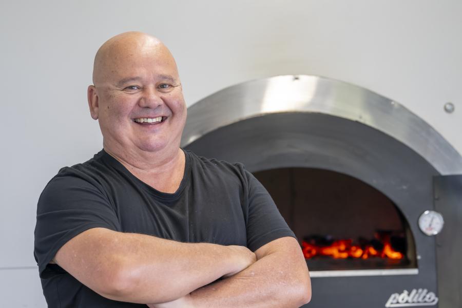 Man smiles with arms folded in front of pizza oven