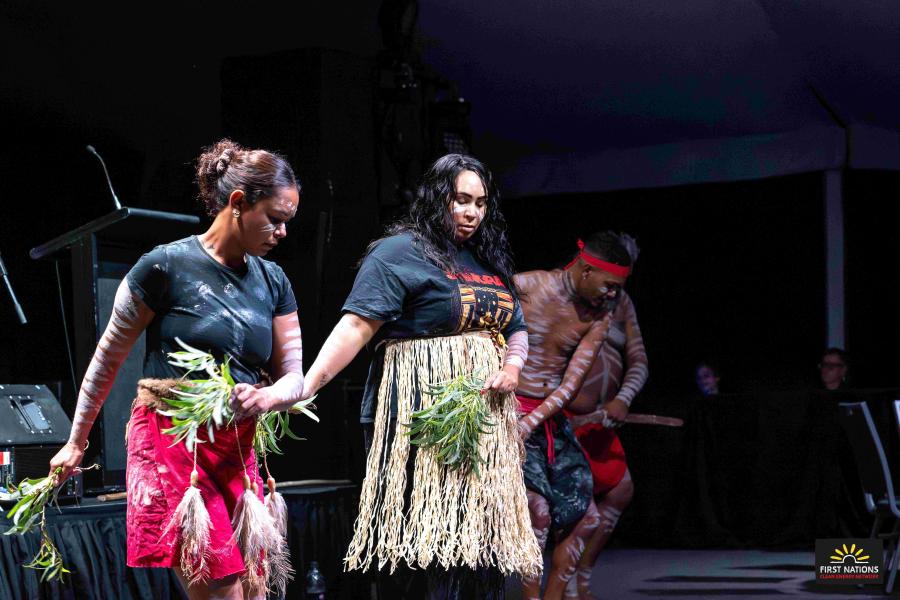 Four First Nations dancers performing in front of a stage and podium.