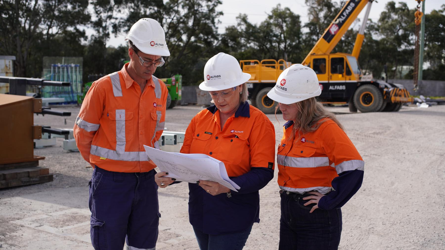 Three people in high-vis safety gear look at a plan in front of construction equipment