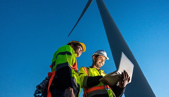 Two workers looking at a laptop in front of a wind turbine