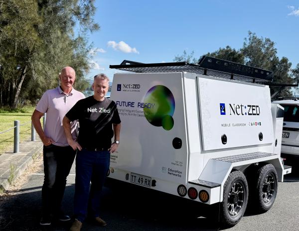 Two men, one in a branded Net Z(ed) tee shirt, standing next to a mobile classroom trailer